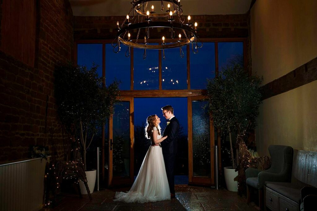 A bride in a flowing white gown and a groom in a dark suit share a romantic moment, standing closely together under a grand chandelier in a warmly lit room with large glass doors revealing a dark blue twilight sky. Image by Documentary Wedding Photographer Lewis Bishop Photography.
