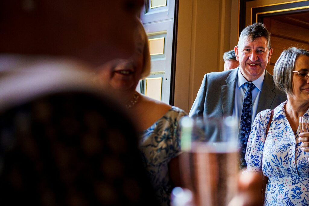 A group of people socialize indoors, chatting and holding drinks. A man in a blue suit and tie smiles warmly in the background. Two women in light-colored dresses are engaged in conversation in the foreground. The setting appears to be bright and lively. Image by Documentary Wedding Photographer Lewis Bishop Photography.