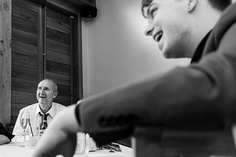 A black and white photo of two men at a table. An older man on the left, wearing a shirt and tie, is smiling and looking toward the younger man on the right, who is also smiling. The younger man is closer to the camera and slightly out of focus. Image by Documentary Wedding Photographer Lewis Bishop Photography.