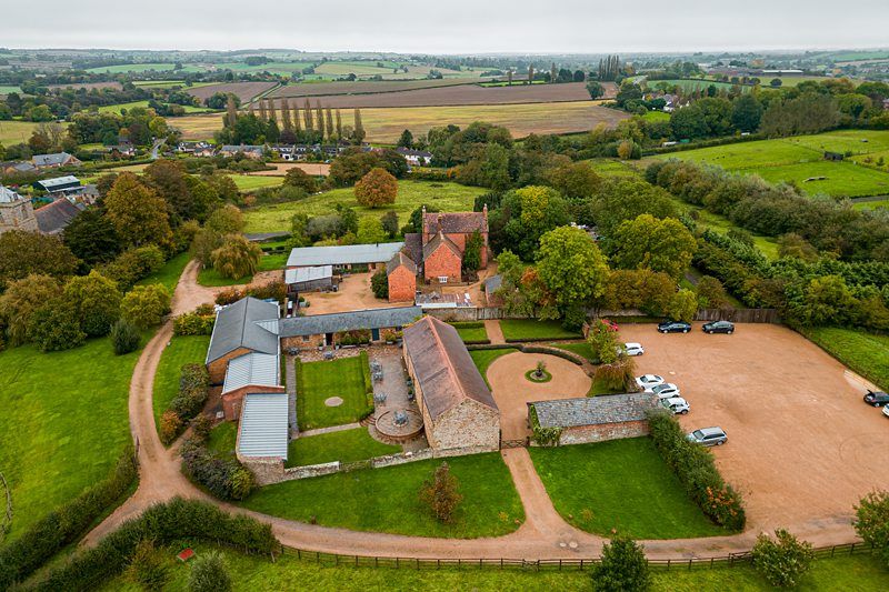 An aerial view of a large countryside estate surrounded by fields and greenery. The estate includes several brick buildings with red roofs, a circular driveway, and a few parked cars. Various trees and a dirt road are visible in the landscape. Image by Documentary Wedding Photographer Lewis Bishop Photography.