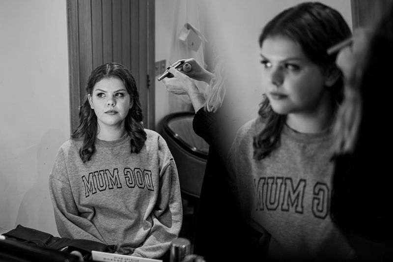 A woman with wavy hair looks at herself in the mirror while having her makeup done. She is wearing a sweatshirt that reads "DOG MUM." The scene is indoors, with a reflection of her face visible in the mirror. Image by Documentary Wedding Photographer Lewis Bishop Photography.