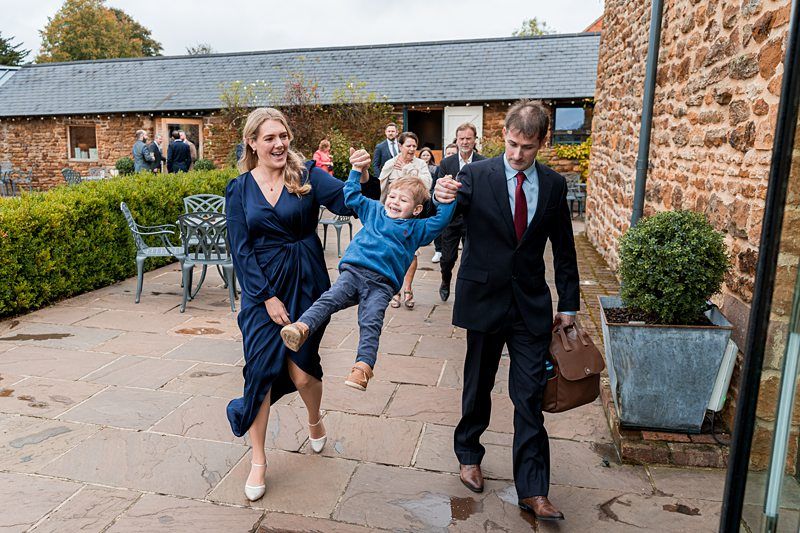 A woman in a blue dress and a man in a suit joyfully swing a smiling child between them as they walk on a stone path. Several other people are in the background near a rustic building with stone walls and a slate roof, all appearing happy and engaged. Image by Documentary Wedding Photographer Lewis Bishop Photography.