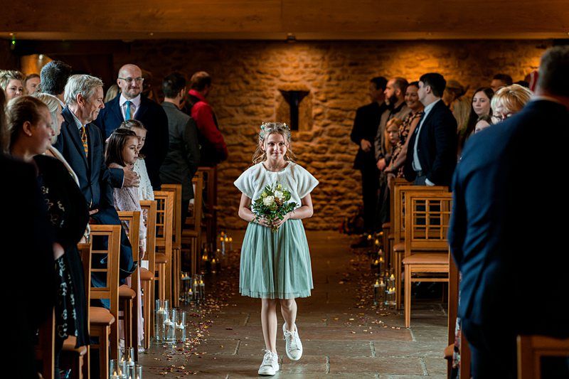 A young girl wearing a green dress, white top, and white shoes walks down the aisle in a stone-walled venue, holding a bouquet of flowers. Guests, dressed in formal attire, are seated on either side, watching her with smiles. Image by Documentary Wedding Photographer Lewis Bishop Photography.