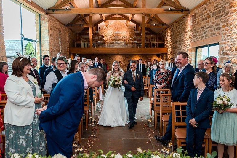 A bride in a white dress, holding a bouquet and escorted by an older man, walks down the aisle of a rustic indoor venue. Guests seated on both sides smile and look on. The groom, dressed in blue, bows his head near the aisle. The space has stone walls and wooden beams. Image by Documentary Wedding Photographer Lewis Bishop Photography.