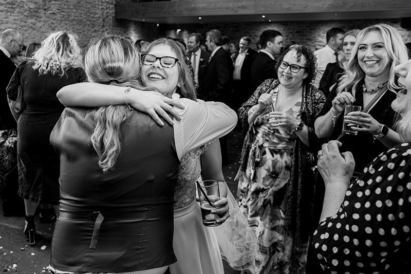 A black and white photo capturing a joyful moment at a gathering. Two women are embracing in the foreground, surrounded by smiling and laughing people holding drinks. The background shows a rustic venue with stone walls and a high ceiling. Image by Documentary Wedding Photographer Lewis Bishop Photography.