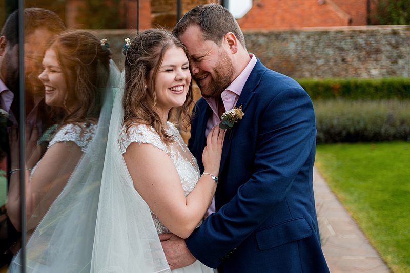A joyful couple on their wedding day stands close together, reflecting on a glass wall. The bride, in a bridal gown and veil, holds the lapel of the groom's blue suit as they smile lovingly at each other. The background features a stone wall, grass, and vibrant greenery. Image by Documentary Wedding Photographer Lewis Bishop Photography.