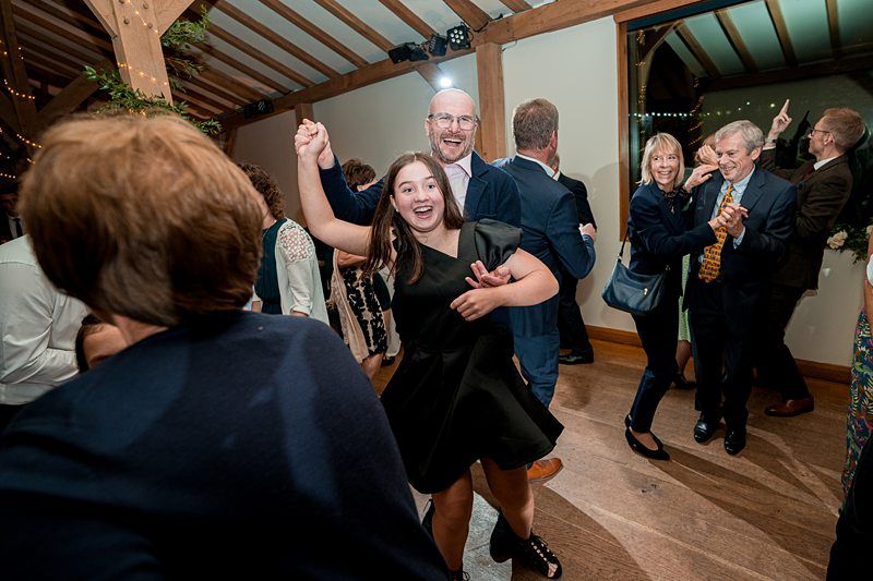 A lively scene at a wedding reception, with a bald man and a young girl dancing energetically in the center. They are surrounded by other guests, who are also dancing and mingling. The venue has wooden beams and festive lighting, creating a warm and joyful atmosphere. Image by Documentary Wedding Photographer Lewis Bishop Photography.