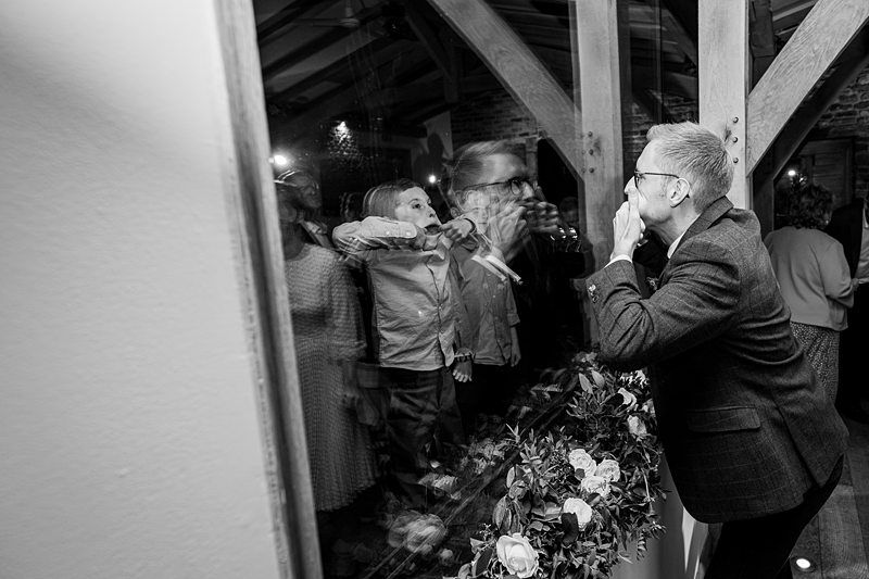 A man in a suit playfully kisses a glass window while a child inside mirrors his action. The scene takes place in a rustic venue with wooden beams and flowers in the foreground. Other people are blurred in the background. The image is in black and white. Image by Documentary Wedding Photographer Lewis Bishop Photography.