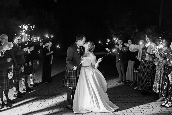 Couple with sparklers, kilt-wearing guests at night wedding.