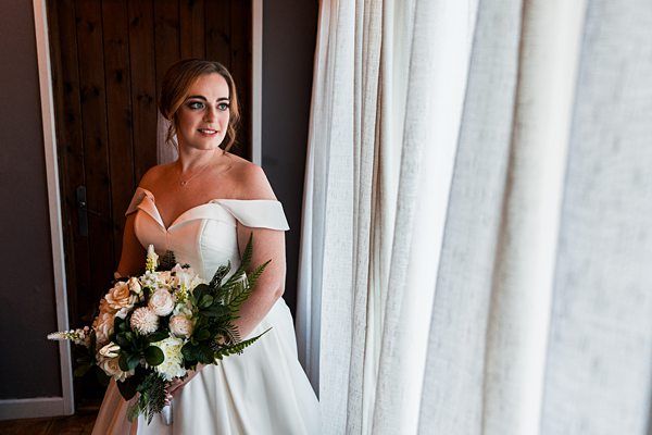 A bride in a white off-shoulder wedding dress holding a bouquet of white and green flowers stands near a window with cream-colored curtains. She gazes out, with a wooden door in the background on the left side. Image by Documentary Wedding Photographer Lewis Bishop Photography.