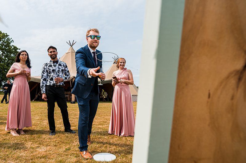 A man in a blue suit and sunglasses is tossing a ring toward a target at an outdoor event. Three people in formal attire, including two women in pink dresses and a man in a patterned shirt, watch and smile. A large tent and trees are visible in the background. Image by Documentary Wedding Photographer Lewis Bishop Photography.