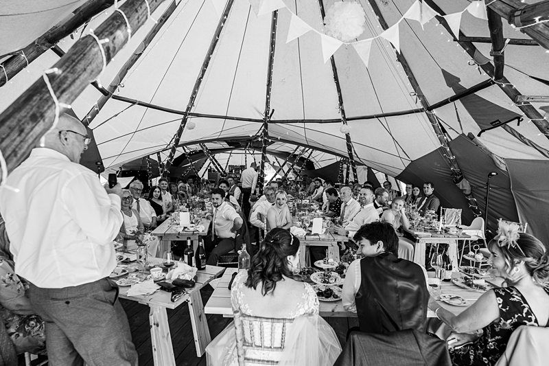 A black and white photo shows a large gathering under a tent. People are seated at tables, enjoying a meal while a person stands at one end, appearing to give a speech. The atmosphere is festive, with decorations like bunting hanging from the tent's poles. Image by Documentary Wedding Photographer Lewis Bishop Photography.