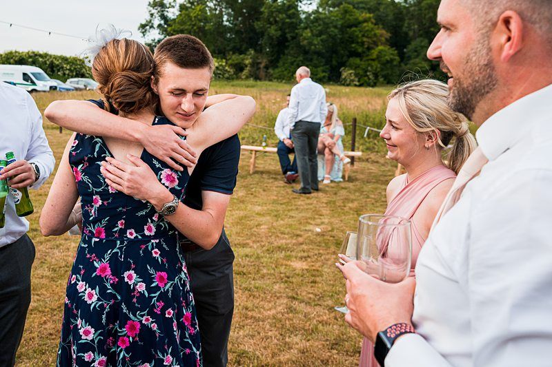 A person in a dark shirt and a person in a floral dress embrace at an outdoor gathering. Two other individuals, one holding two glasses, stand nearby watching. In the background, more people are seated on benches with trees and parked cars visible. Image by Documentary Wedding Photographer Lewis Bishop Photography.