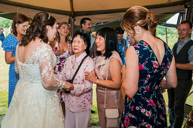 A bride in a white lace wedding dress greets an older woman in a light pink ensemble under a canopy. They are surrounded by smiling guests, dressed in colorful attire, engaging in conversation and laughter. The atmosphere is festive and celebratory. Image by Documentary Wedding Photographer Lewis Bishop Photography.