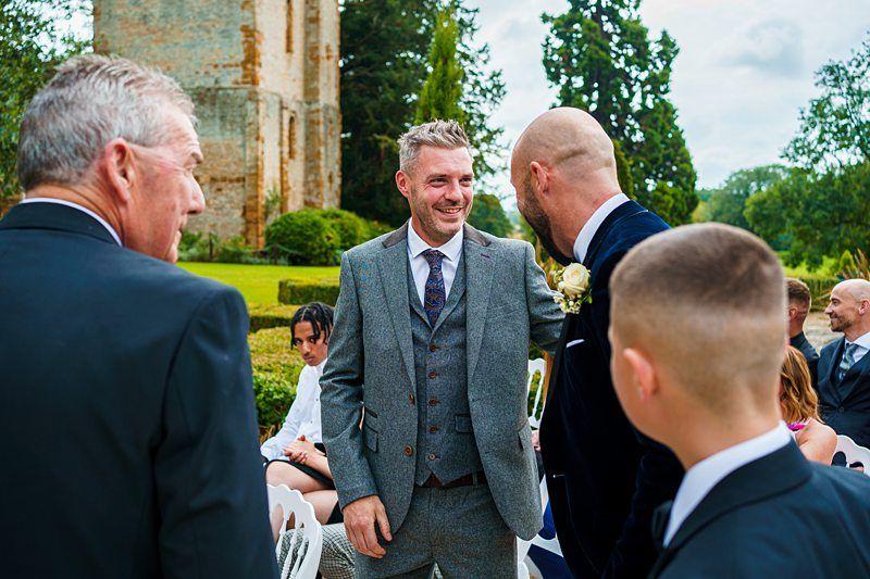A group of men in suits, one of whom smiles and greets another with a friendly hand on the shoulder, in an outdoor setting with an old stone structure and trees in the background. Other people are seated around, suggesting a formal event like a wedding. Image by Documentary Wedding Photographer Lewis Bishop Photography.