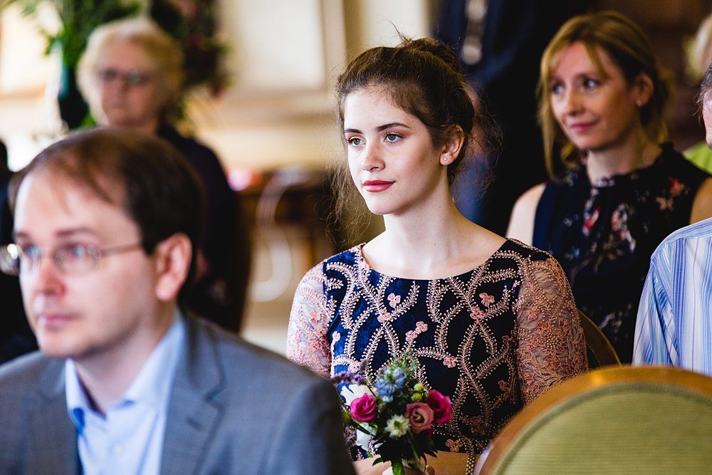 A young woman with brown hair tied back is holding a small bouquet of flowers while sitting in an audience. She is wearing a patterned dress and looking forward. Other blurred attendees are seated around her in the background. Image by Documentary Wedding Photographer Lewis Bishop Photography.