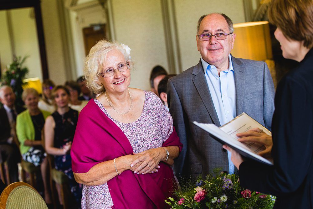 An elderly couple stands together during a ceremony, smiling as they listen to the officiant holding a booklet. The woman is wearing a pink shawl over a floral dress, and the man is in a gray suit. People are seated in the background, watching attentively. Image by Documentary Wedding Photographer Lewis Bishop Photography.