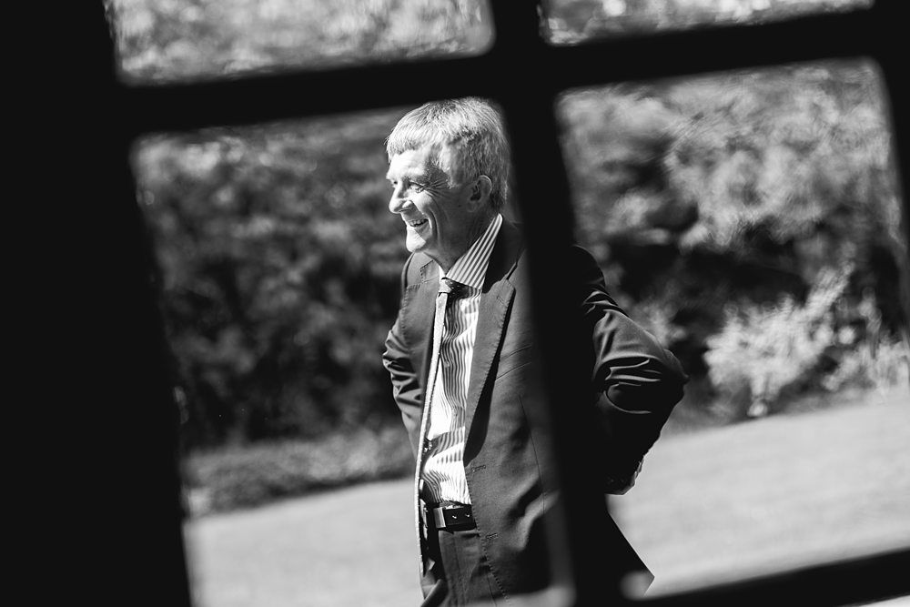 Black and white photo of an older man smiling and looking to the side. He is wearing a suit and standing outdoors in a garden. The photo is framed through a grid-like window, adding a unique perspective. Trees and foliage are visible in the blurred background. Image by Documentary Wedding Photographer Lewis Bishop Photography.
