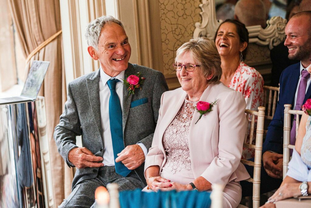 An older man in a grey suit with a blue tie and an older woman in a light pink outfit with a matching floral corsage sit together and smile at a formal event. Behind them, other guests are engaged and smiling, seated on wooden chairs in a decorated room. Image by Documentary Wedding Photographer Lewis Bishop Photography.