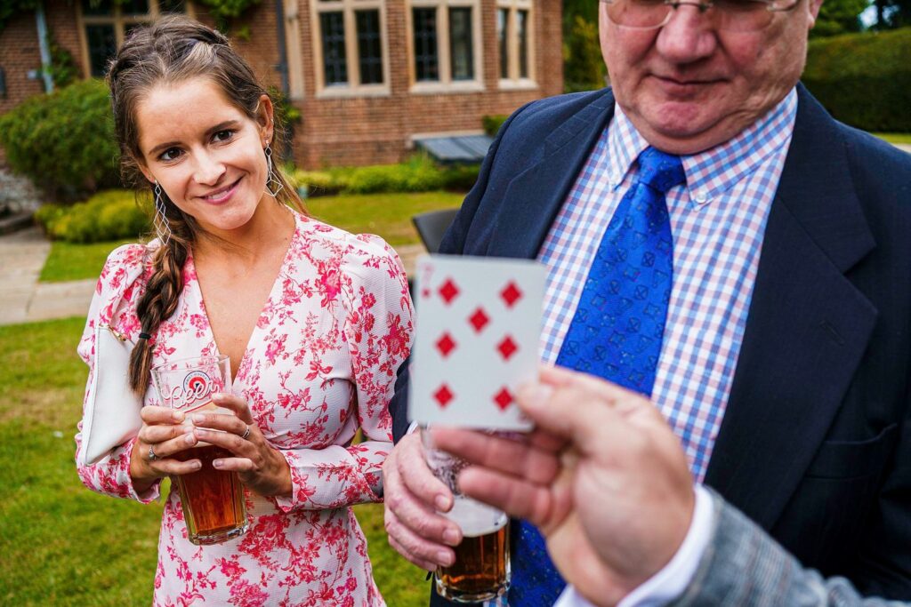 A woman in a floral dress and a man in a suit watch a person holding a six of diamonds playing card. The woman smiles while holding a glass of beer. They appear to be outdoors in front of a brick building with windows. Image by Documentary Wedding Photographer Lewis Bishop Photography.
