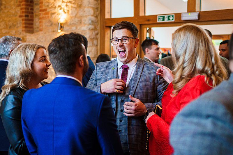A man in a gray checkered suit and glasses is laughing and talking with three other people at a social event. One woman has blonde hair and wears a red outfit. The background features a stone wall and large windows, creating a warm and inviting atmosphere. Image by Documentary Wedding Photographer Lewis Bishop Photography.
