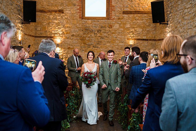 A smiling couple walks hand-in-hand down an aisle as newlyweds. The bride is in a white gown holding a bouquet of red and green flowers, while the groom wears a gray suit. They are surrounded by cheering guests in formal attire, inside a rustic brick venue. Image by Documentary Wedding Photographer Lewis Bishop Photography.