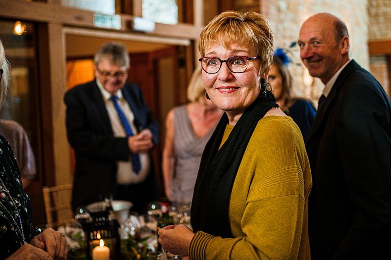 A woman in glasses and a mustard-colored top with a black scarf smiles at a gathering. She is surrounded by other people who are casually dressed, and they appear to be enjoying a social event. Lit candles and a decorated table are visible in the foreground. Image by Documentary Wedding Photographer Lewis Bishop Photography.