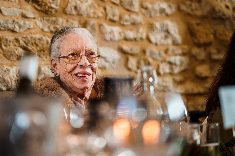 An elderly woman with glasses smiles while seated at a table. She is wearing a fur-lined coat and is surrounded by blurred wine glasses and candles, with a stone wall in the background. The setting appears warm and festive. Image by Documentary Wedding Photographer Lewis Bishop Photography.