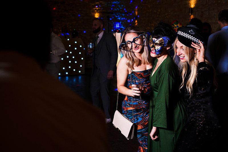 Three women in colorful outfits and costume accessories pose together at a party. The woman on the left wears oversized glasses, the middle one has a mask, and the right one wears a police hat. Party lights and other guests are visible in the background. Image by Documentary Wedding Photographer Lewis Bishop Photography.
