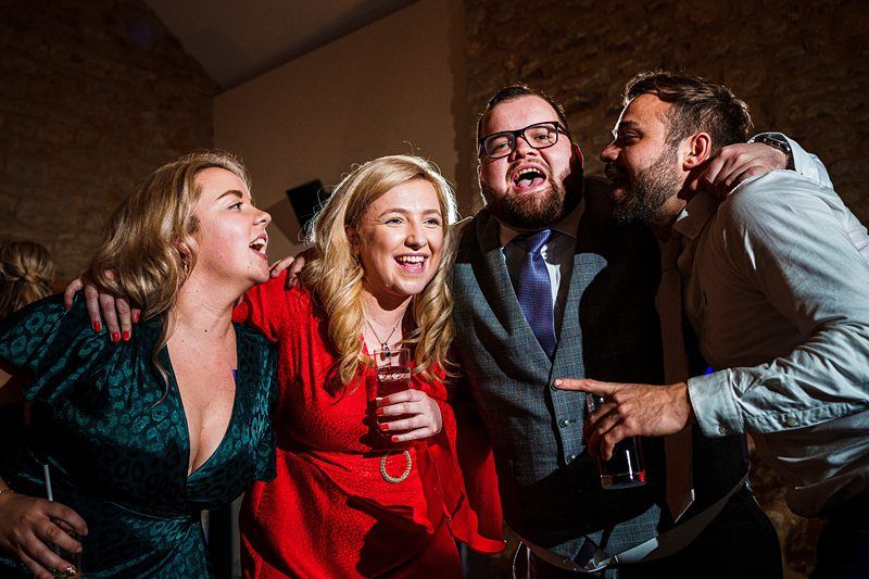 A group of four people are celebrating indoors. Two women in dresses and two men in suits are smiling and have their arms around each other. They appear to be at a party or a wedding reception, and one woman is holding a drink. The background has a stone wall. Image by Documentary Wedding Photographer Lewis Bishop Photography.