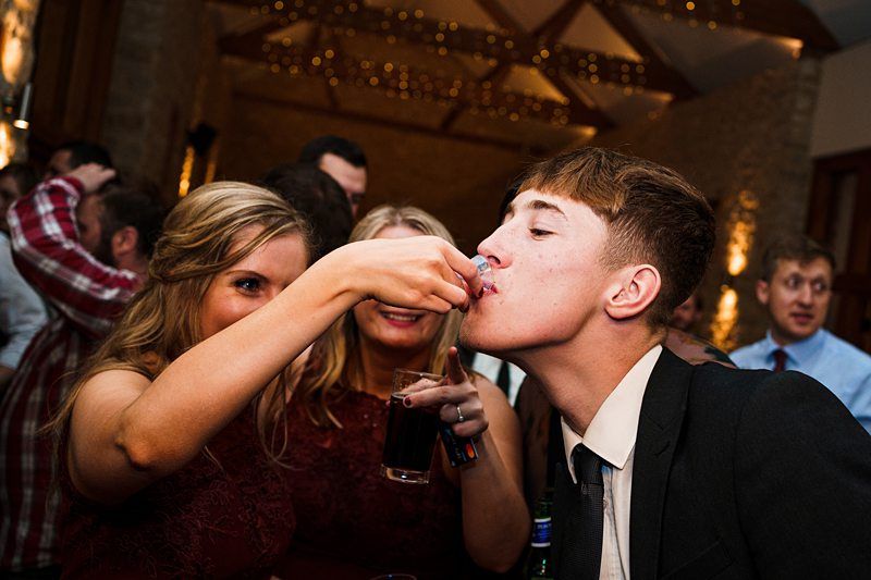 A young man in a suit leans forward to take a shot from a woman’s hand at a lively indoor party. Two women, one holding the shot and another with a drink, smile in the background. Warm lighting and festive string lights create an inviting atmosphere. Image by Documentary Wedding Photographer Lewis Bishop Photography.