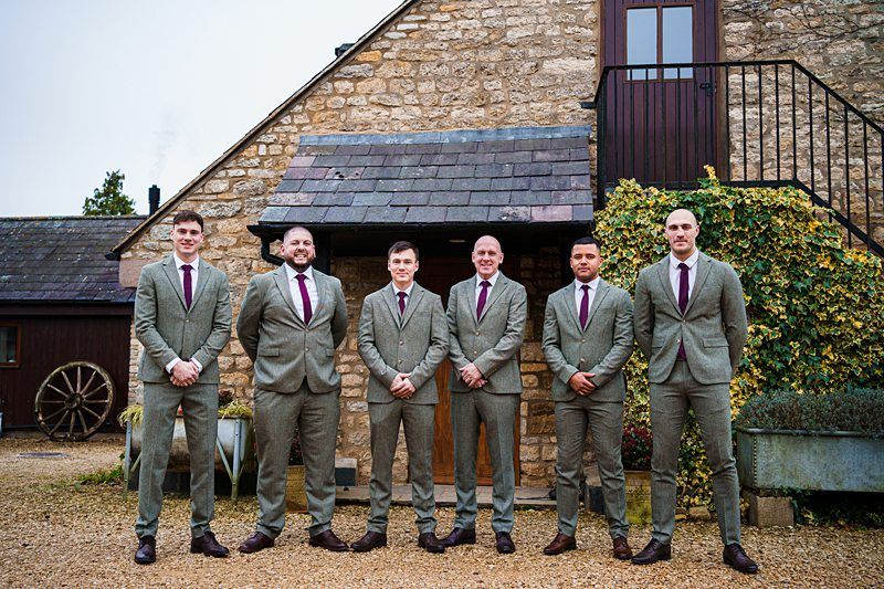 Six men stand in front of a rustic stone building with stairs leading to a door. They are dressed in matching grey suits, white shirts, and burgundy ties. Behind them, there are plants and a decorative wagon wheel. The setting appears to be outdoors. Image by Documentary Wedding Photographer Lewis Bishop Photography.