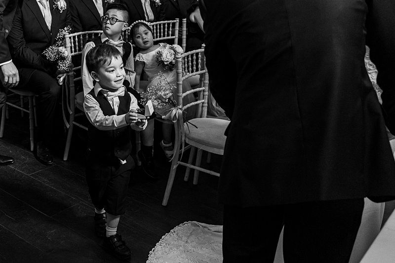 A young boy, dressed in formal attire, walks down the aisle holding a box, possibly carrying rings. He is watched by seated guests, some of whom are children also in formal wear. The scene appears to be at a wedding ceremony, captured in black and white. Image by Documentary Wedding Photographer Lewis Bishop Photography.