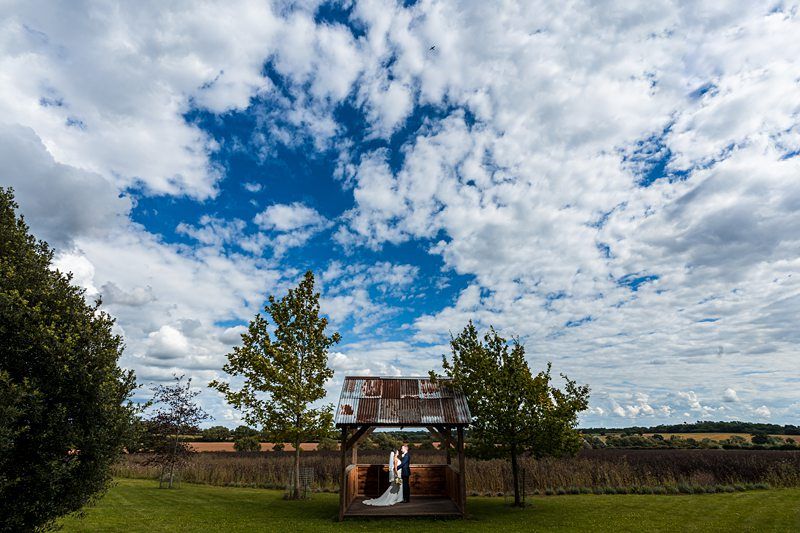 A newlywed couple stands under a rustic wooden shelter on a vast, grassy field. The sky above is a vibrant blue, dotted with fluffy white clouds. Trees and open landscape stretch out in the background, creating a serene and picturesque setting. Image by Documentary Wedding Photographer Lewis Bishop Photography.