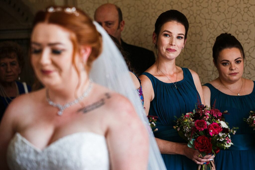 A bride in a white dress and veil stands in the foreground, slightly out of focus. Behind her are two bridesmaids in teal dresses, one of whom holds a bouquet of red and white flowers. Several other people are partially visible in the background. Image by Documentary Wedding Photographer Lewis Bishop Photography.