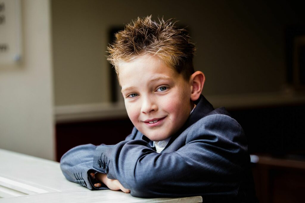 A young boy with spiky brown hair, dressed in a dark blue suit and white shirt, leans his arms on a white surface. He is smiling softly and looking directly at the camera. The background is blurred and dark, putting emphasis on the boy. Image by Documentary Wedding Photographer Lewis Bishop Photography.