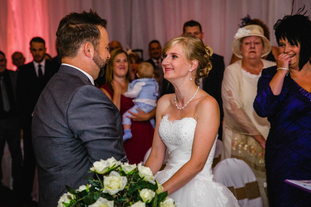A bride and groom stand facing each other, holding hands and smiling, surrounded by guests at a wedding ceremony. The bride is in a white dress with a bouquet of white roses, and the groom is in a gray suit. People in the background are watching, smiling, and taking photos. Image by Documentary Wedding Photographer Lewis Bishop Photography.
