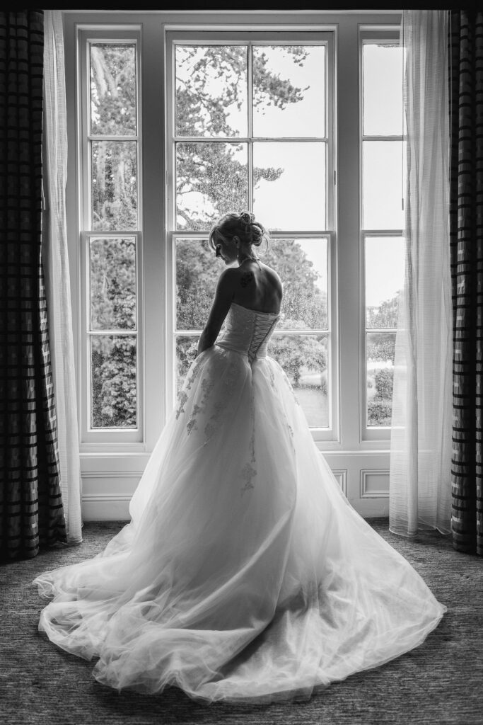 Black-and-white photo of a woman in a wedding dress standing in front of a large window. The skirt of her dress flows out behind her, and she gazes downward. The window offers a view of trees and greenery outside. Drapes frame the scene on either side. Image by Documentary Wedding Photographer Lewis Bishop Photography.