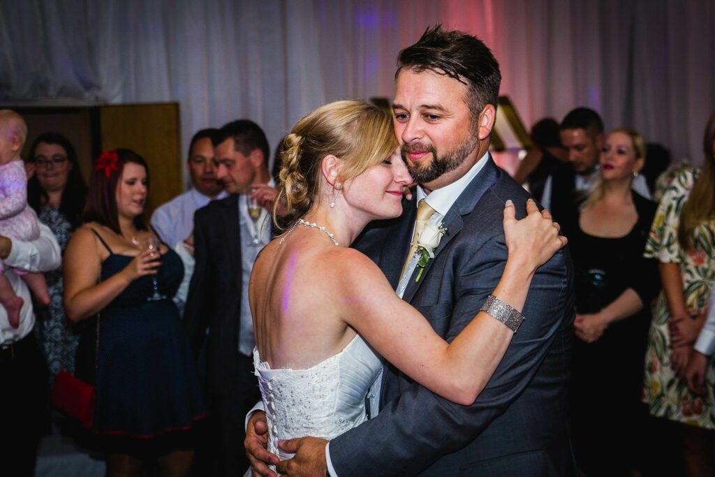 A bride and groom share a close embrace while dancing at their wedding reception. The bride wears a white lace dress, and the groom is in a gray suit with a white tie. Guests, some with drinks and others watching, surround them. A baby is held on the left side. Image by Documentary Wedding Photographer Lewis Bishop Photography.