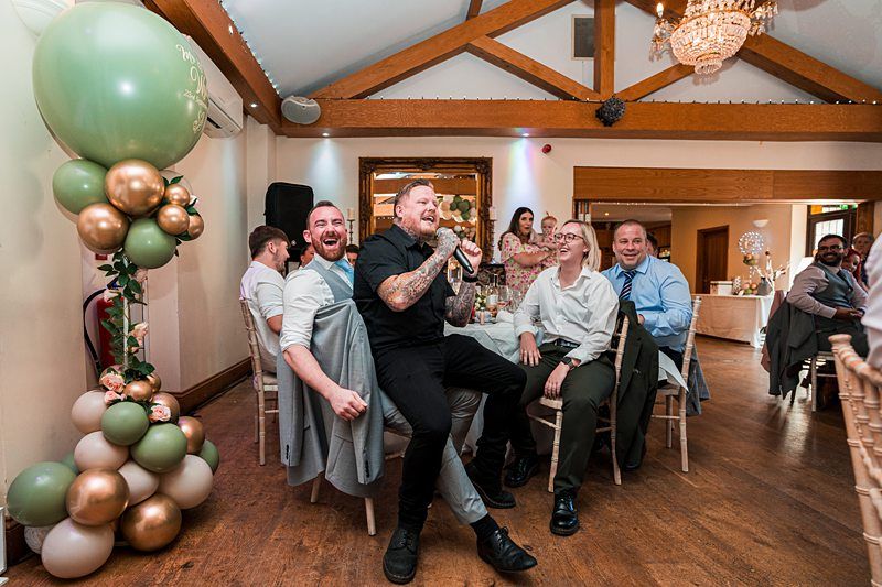 A group of people enjoy a lively moment at a celebration. One man, holding a microphone, sings passionately while seated among his smiling friends at a decorated venue. Green and gold balloons are visible in the foreground, with chandeliers hanging from the ceiling. Image by Documentary Wedding Photographer Lewis Bishop Photography.