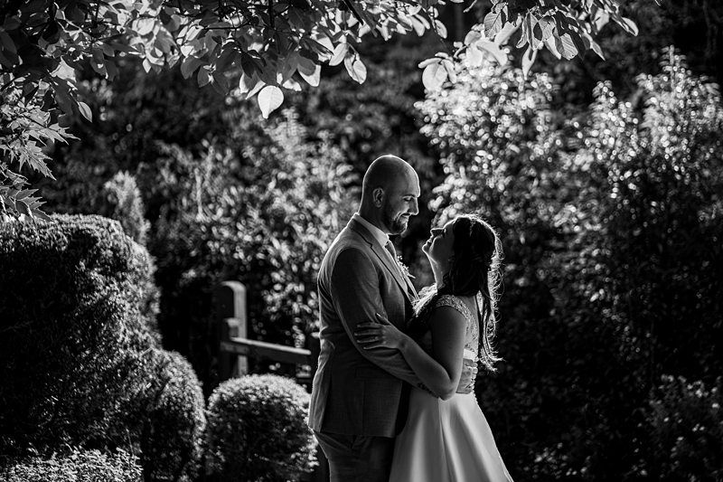 A black and white photo of a couple on their wedding day, standing outdoors, surrounded by lush foliage. The groom is wearing a suit and the bride is in a flowing wedding dress. They are gazing into each other's eyes and smiling warmly. Image by Documentary Wedding Photographer Lewis Bishop Photography.