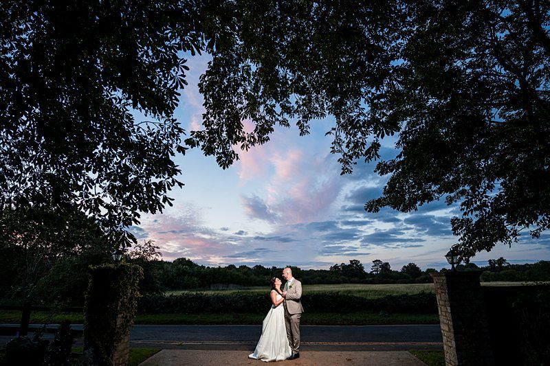 A newlywed couple embraces and poses in a picturesque outdoor setting at dusk. The sky is painted with hues of pink and blue, framed by silhouetted trees, adding a romantic backdrop to the scene. The bride is in a white dress, and the groom is in a light-colored suit. Image by Documentary Wedding Photographer Lewis Bishop Photography.