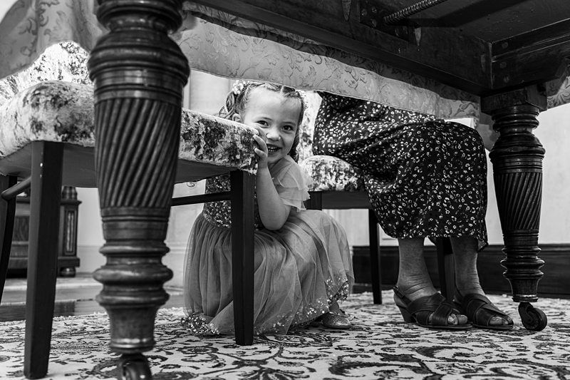 A little girl in a dress crouches under a table next to a woman sitting in a chair. The girl is smiling and looking at the camera. The scene is in black and white with ornate furniture and a patterned rug visible. Image by Documentary Wedding Photographer Lewis Bishop Photography.