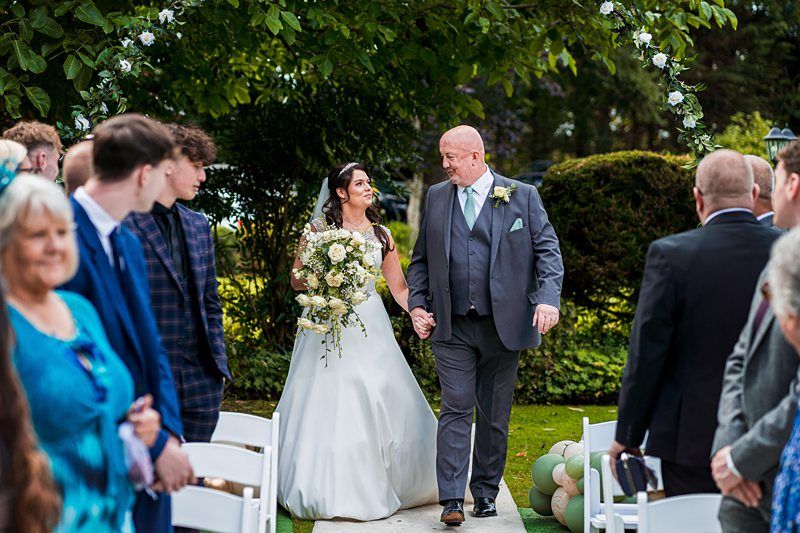 A bride, in a white gown holding a bouquet, walks down an outdoor aisle with a man in a gray suit. Guests, dressed formally, are seated on either side. The aisle is adorned with greenery and white flowers, and trees and bushes are visible in the background. Image by Documentary Wedding Photographer Lewis Bishop Photography.