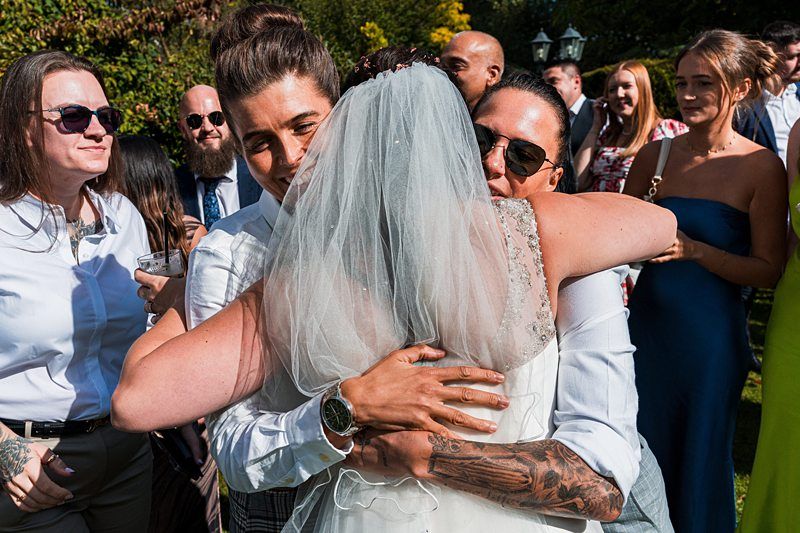 A bride in a white dress and veil is hugging two people in a sunny outdoor setting, surrounded by smiling guests. One guest holds a drink, while others are dressed in semi-formal attire, enjoying what appears to be a celebratory event. Image by Documentary Wedding Photographer Lewis Bishop Photography.