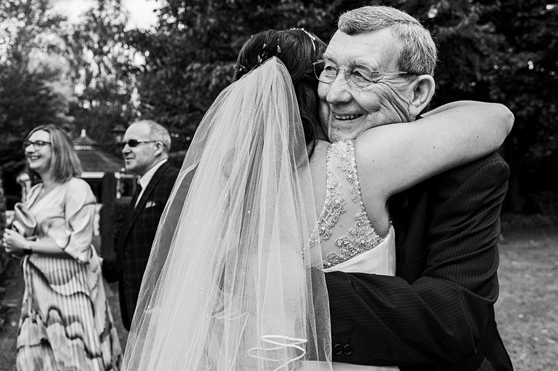 A joyful newlywed, wearing a veil, embraces an elderly man in a heartfelt hug. The joyous moment is captured in a black and white photograph, with two other guests smiling and chatting in the background, highlighting the festive atmosphere of the outdoor event. Image by Documentary Wedding Photographer Lewis Bishop Photography.