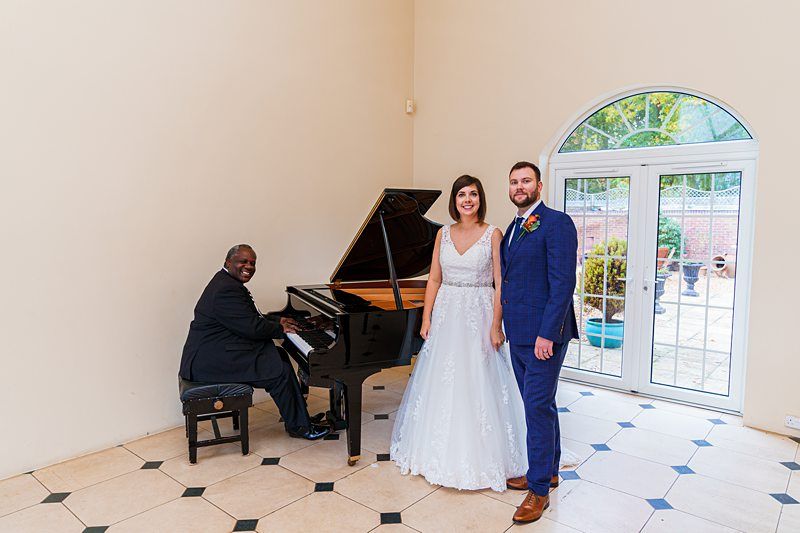A couple dressed in wedding attire stands indoors next to a grand piano. The groom is in a blue suit and the bride in a white gown. A man in a black suit is seated at the piano, smiling and playing. They are all in a bright room with large windows behind them. Image by Documentary Wedding Photographer Lewis Bishop Photography.