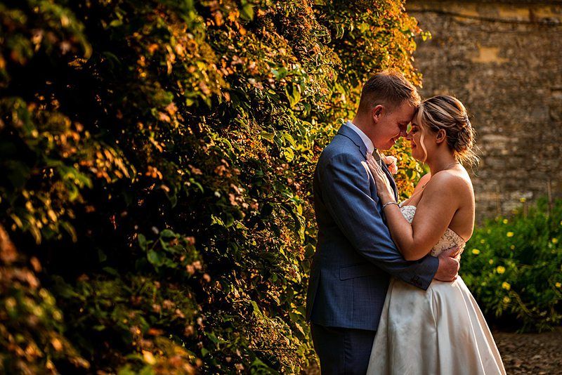 A couple dressed in wedding attire embraces closely in an intimate moment. They stand beside a sunlit, leafy wall. The groom in a light blue suit gently holds the bride in a white gown, their foreheads touching as they share a loving gaze. Image by Documentary Wedding Photographer Lewis Bishop Photography.