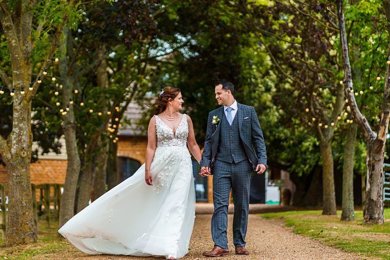 A bride and groom walk hand-in-hand down a tree-lined path adorned with string lights. The bride wears a flowing white gown, and the groom is in a checked suit. They are smiling at each other, surrounded by lush greenery, creating a romantic atmosphere. Image by Documentary Wedding Photographer Lewis Bishop Photography.