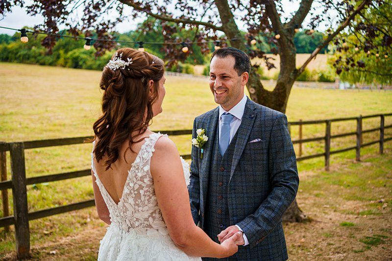 A bride and groom stand outdoors, holding hands and smiling at each other. The bride wears a white lace dress with her hair styled in waves. The groom is dressed in a checkered suit and light blue tie. They are under a tree with string lights, with a field in the background. Image by Documentary Wedding Photographer Lewis Bishop Photography.